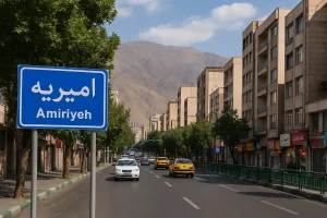 تابلوی آبی رنگ «امیریه» در کنار خیابان اصلی محله امیریه تهران دیده میشود؛ خیابانی خلوت با ساختمانهای چندطبقه، درختان دو طرف مسیر و نمای کوههای البرز در پسزمینه زیر آسمان صاف و آفتابی.