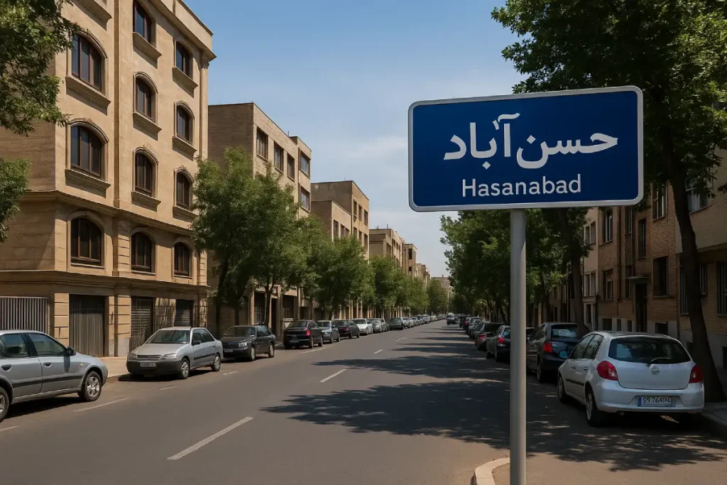 نمایی از خیابانی آرام در محله حسن‌آباد با ساختمان‌های آجری و سنگی در دو طرف، درختان سبز، خودروهای پارک‌شده در حاشیه خیابان و تابلوی آبی‌رنگی که نام «حسن‌آباد» روی آن نوشته شده است.
