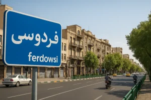 نمایی از خیابان فردوسی تهران با ساختمان‌های تاریخی در دو طرف خیابان، درختان سبز، خودروها و موتورسیکلت‌ها در حال حرکت در روزی آفتابی.