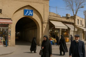 ورودی سنتی بازار با طاق آجری و تابلوی آبی‌رنگ «بازار» در بالای درگاه، افرادی در حال عبور از مقابل مغازه‌های قدیمی با اجناس رنگارنگ، در فضایی گرم و روزمره از بافت تاریخی شهر.