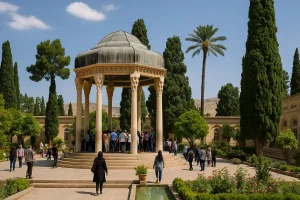 نمایی از آرامگاه حافظ در شیراز، با عمارت هشت‌ضلعی و گنبد فیروزه‌ای در میان درختان سرو و نخل بلند، بازدیدکنندگان در اطراف آرامگاه در حال قدم‌زدن و تماشای فضای سرسبز و آرام محوطه هستند.