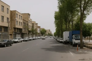 نمایی از خیابانی آرام در محله فروزش با ساختمانهای چندطبقه آجری، خودروهای پارکشده در دو طرف خیابان، و درختان بلند که سایهای دلنشین روی مسیر ایجاد کردهاند در یک روز نیمهابری.