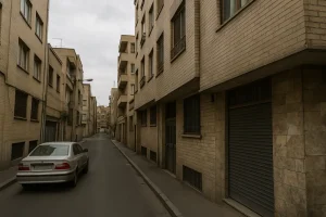 نمایی از کوچه‌ای باریک و آرام با ساختمان‌های آجری در دو طرف، پنجره‌های فلزی و یک خودروی نقره‌ای پارک‌شده در کنار خیابان زیر آسمانی ابری.