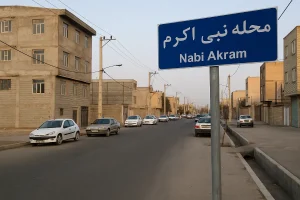 نمایی از محله نبی اکرم با خیابانی آرام، ساختمانهای مسکونی آجری و چند خودرو پارکشده در کنار خیابان در یک روز صاف و آفتابی.