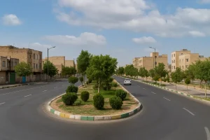 خیابانی خلوت در محله خاوران با ساختمانهای آجری کمارتفاع در دو طرف، درختان سبز در فضای سبز وسط خیابان و آسمانی آبی با ابرهای پراکنده در روزی آفتابی.