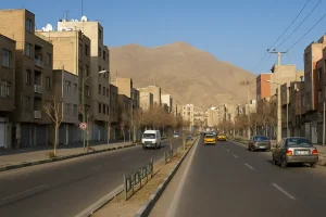 نمایی از محله آهنگ تهران، خیابان دوطرفه با ساختمانهای آجری چندطبقه، خودروهای در حال حرکت و کوه در پسزمینه در یک روز آفتابی"