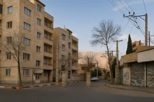 خیابان آرامی در یک محله مسکونی ایران، با ساختمان‌های چندطبقه سنگی به رنگ کرم، درختان خشکیده زمستانی، خودروهای پارک‌شده و کوه‌های روشن در پس‌زمینه زیر آسمان آبی صاف.