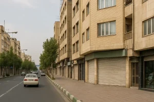 «نمایی از یک خیابان خلوت در محلهای شهری با ساختمانهای چندطبقه آجری، مغازههای بسته، چند خودرو در حال حرکت و درختهای ردیفی در دو طرف خیابان.»