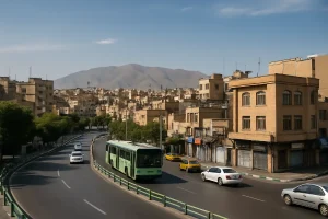 «نمایی از خیابانی در محلهای قدیمی در تهران با ساختمانهای آجری متعدد، اتوبوس سبز در حال حرکت در مسیر اصلی، چند خودروی سواری، درختان کنار خیابان و رشتهکوههای بلند در پسزمینه زیر آسمان صاف.»