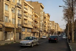 «نمایی از یک خیابان مسکونی در شهری ایرانی، با ساختمانهای چندطبقه، خودروهای در حال عبور و درختان خشک در دو طرف خیابان در نور زرد غروب.»