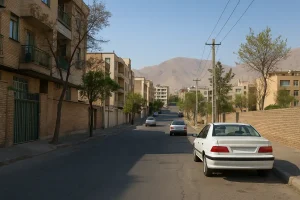 «نمایی واقعی از یک خیابان مسکونی آرام با ساختمانهای آجری چندطبقه، چند خودرو پارکشده در کنار خیابان، درختان پراکنده و کوههای بلند در پسزمینه زیر آسمان صاف و آفتابی.»