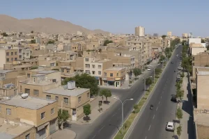 «نمایی هوایی از محلهای قدیمی با بافت فشرده و ساختمانهای کوتاهِ آجریرنگ، خیابان عریض و خلوت در سمت راست تصویر، درختهای پراکنده کنار خیابان و کوهستان خشک در پسزمینه زیر آسمان صاف و آبی.»