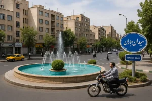 «نمایی واقعگرایانه از میدان بهاران، شامل یک فواره دایرهای با آبِ آبی، ساختمانهای مسکونی اطراف، درختان حاشیه خیابان و عبور یک موتورسوار از کنار میدان در روزی آفتابی.»