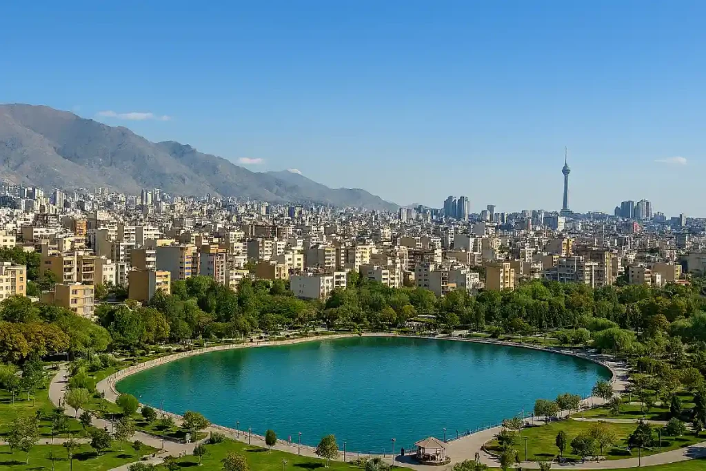 «نمایی پانوراما از محله‌ای شهری در تهران با دریاچه‌ای مصنوعی در مرکز پارک، ساختمان‌های مسکونی متراکم در پس‌زمینه، درختان سرسبز در اطراف، و برج میلاد که در دوردست دیده می‌شود.»