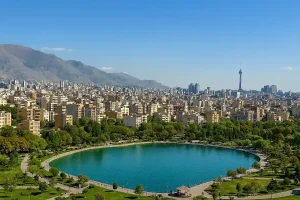 «نمایی پانوراما از محلهای شهری در تهران با دریاچهای مصنوعی در مرکز پارک، ساختمانهای مسکونی متراکم در پسزمینه، درختان سرسبز در اطراف، و برج میلاد که در دوردست دیده میشود.»