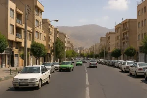 نمایی واقعی از خیابانی در محلهای جنوب تهران با ساختمانهای آجری چندطبقه، درختان کنار خیابان، خودروهای پارکشده و حرکت چند ماشین در مسیر اصلی، در پسزمینه کوههای جنوبی تهران دیده میشود.