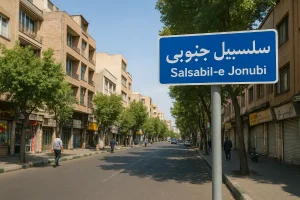 نمایی از خیابان اصلی محله سلسبیل جنوبی در روز، با ساختمانهای آجری و مغازههای قدیمی در دو طرف خیابان، چند عابر پیاده، خودروهای پارکشده و تابلوی آبیرنگی که نام «سلسبیل جنوبی» روی آن نوشته شده است.